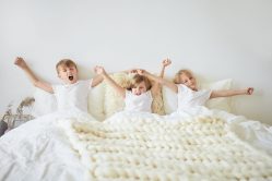 Rise And Shine Isolated Horizontal Shot Of Three Siblings Little Sister And Her Two Elder Brothers Wearing Identical White T Shirts Sitting On Bed Stretching Arms And Yawning In The Morning Min